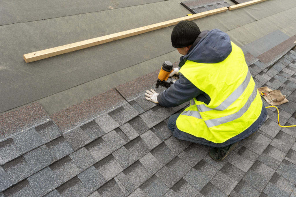 qualified workman in uniform work wear using air or pneumatic nail gun and installing asphalt or bitumen shingle on top of the new roof under construction residential building